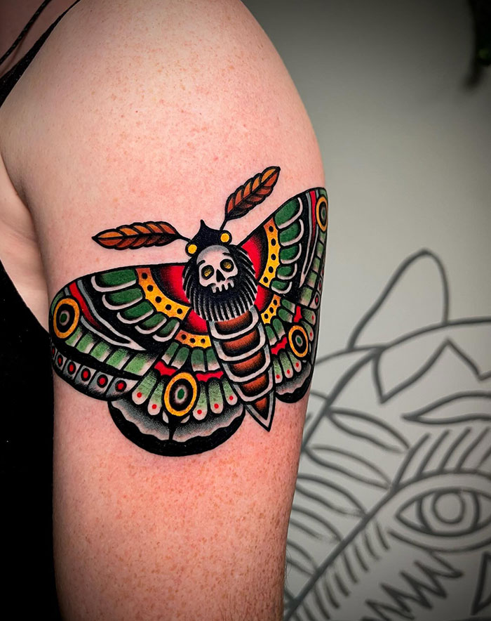 A vibrant tattoo of a moth with colorful wings and a skull on its body, done in traditional American style, on the upper arm.