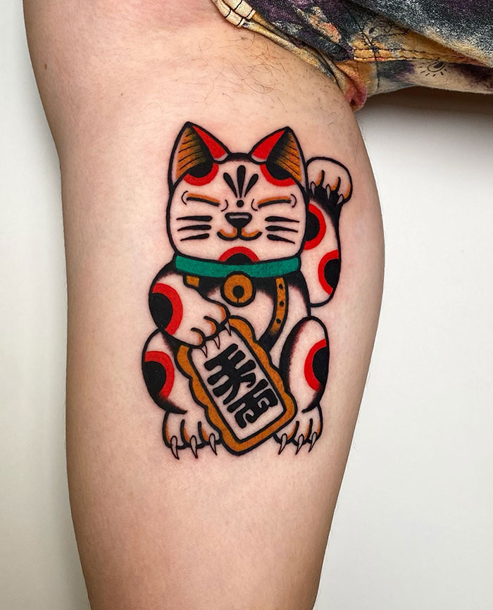 A tattoo of a traditional Japanese lucky cat (Maneki Neko) with red spots, holding a coin and raising one paw, done in bold colors on the thigh.