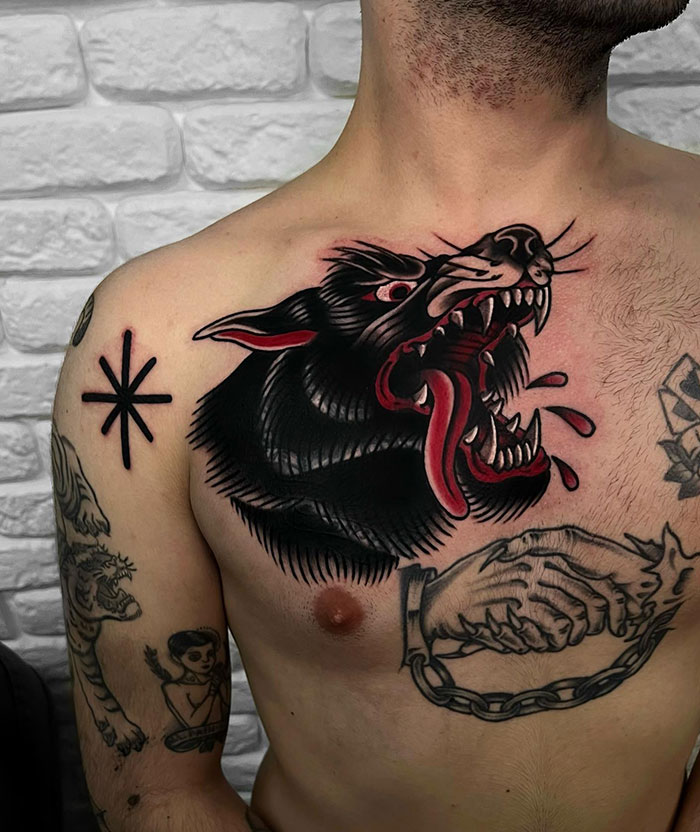 A bold tattoo of a snarling black wolf head with red accents on the chest, featuring detailed shading and blood drops, in traditional style.