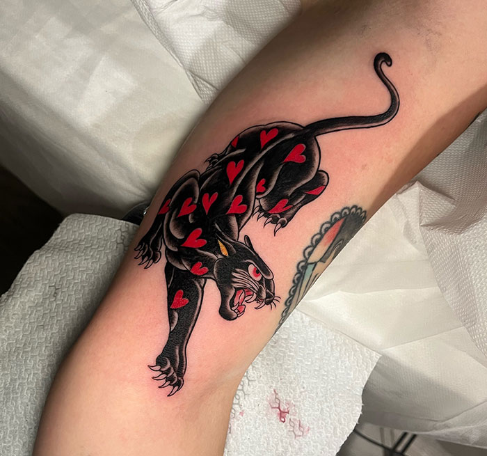 A tattoo of a black panther with red hearts on its body, crouching in a traditional style, featuring bold outlines and vibrant red accents.