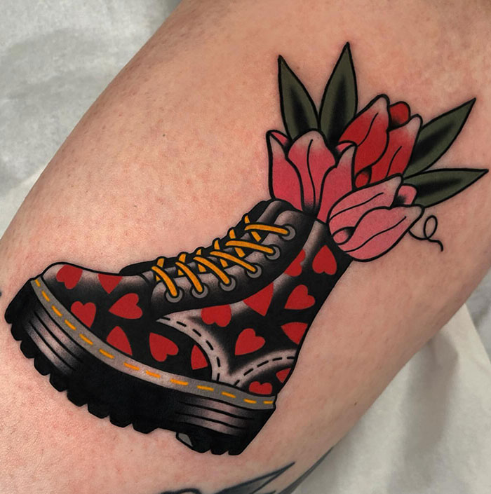 A tattoo of a black boot decorated with red hearts, with pink tulips and green leaves emerging from the top, done in bold traditional style.