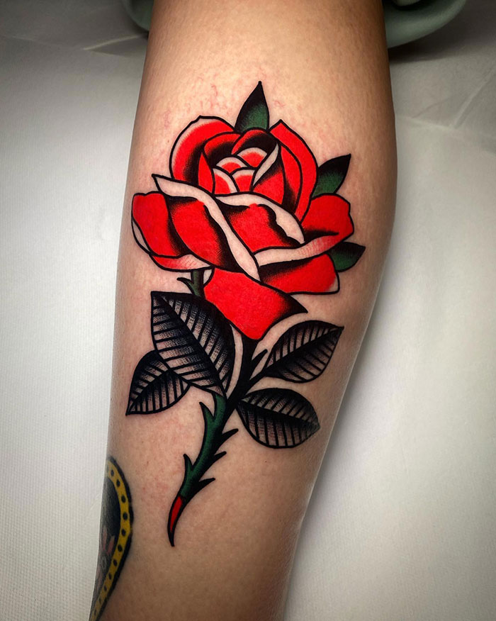 A tattoo of a red rose with detailed petals and bold black shading on the stem and leaves, in traditional American tattoo style on the forearm.