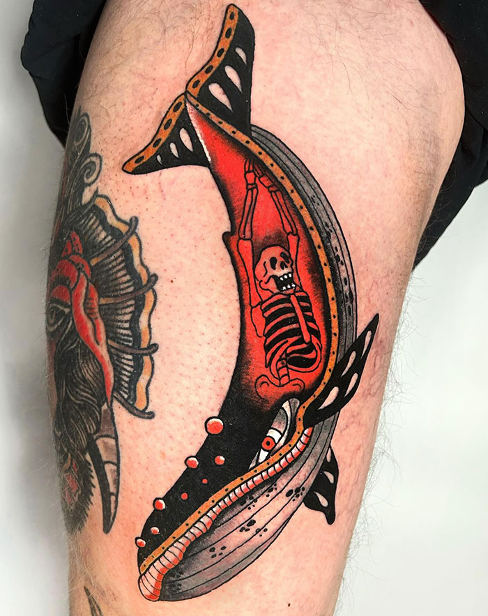 A tattoo of a whale with a visible skeleton inside, filled with red and black shading, done in bold traditional style on the thigh.