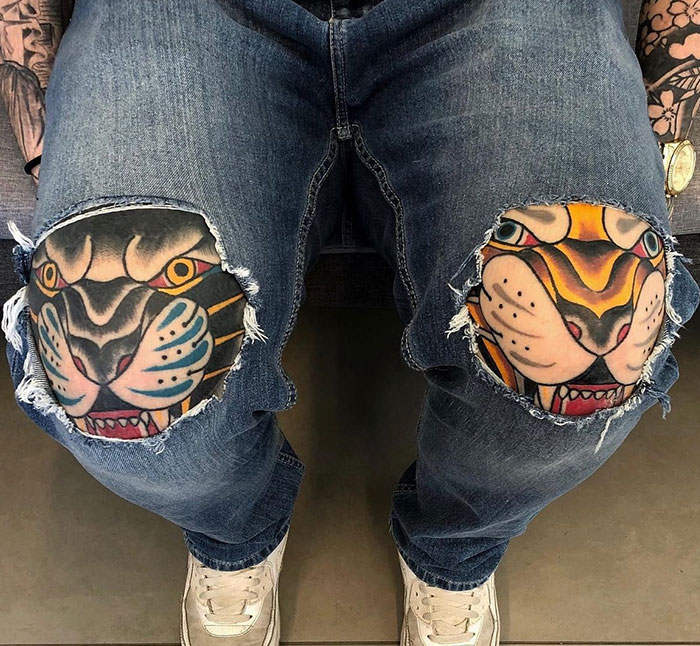 A pair of jeans with ripped knees reveals colorful tiger tattoos on each knee, styled in traditional American tattoo art with bold lines.