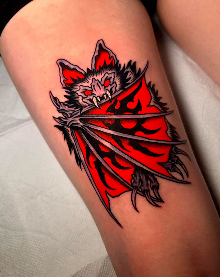 A bold tattoo of a bat with red wings spread wide, featuring traditional American tattoo style with vibrant colors and sharp black outlines.