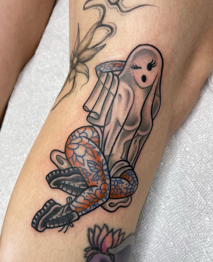 A tattoo of a stylized, feminine ghost wearing patterned stockings and boots, with delicate shading and intricate floral details on the leg.