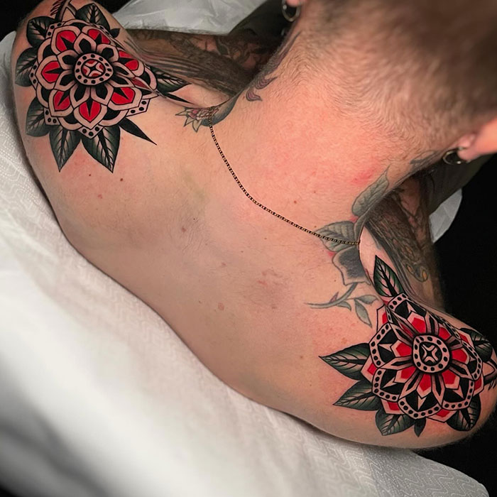 Symmetrical floral tattoos on each shoulder blade, featuring bold red, black, and green traditional American tattoo designs with intricate details.