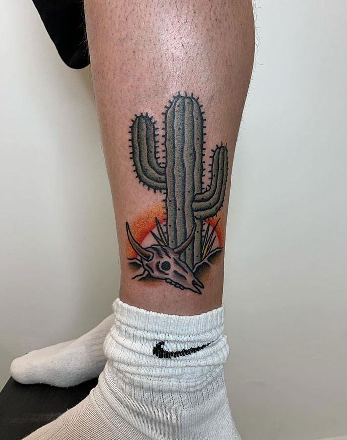 A tattoo of a desert scene with a large cactus, animal skull, and orange sunset in traditional American style on the back of a person's leg.