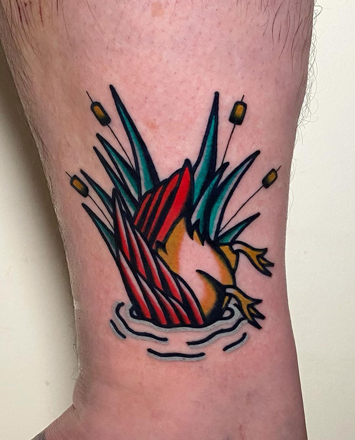 A colorful tattoo of a duck diving underwater with only its tail and legs visible, surrounded by cattails and ripples, in traditional American style.