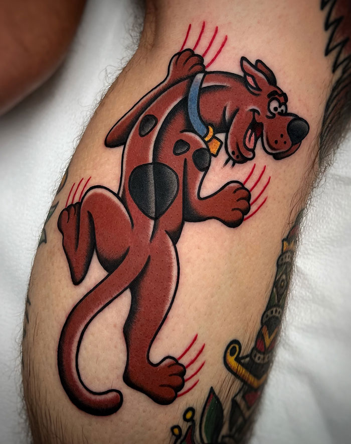 A tattoo of a cartoon dog with a blue collar, scratching at the skin with red claw marks trailing behind, styled in bold colors and outlines.