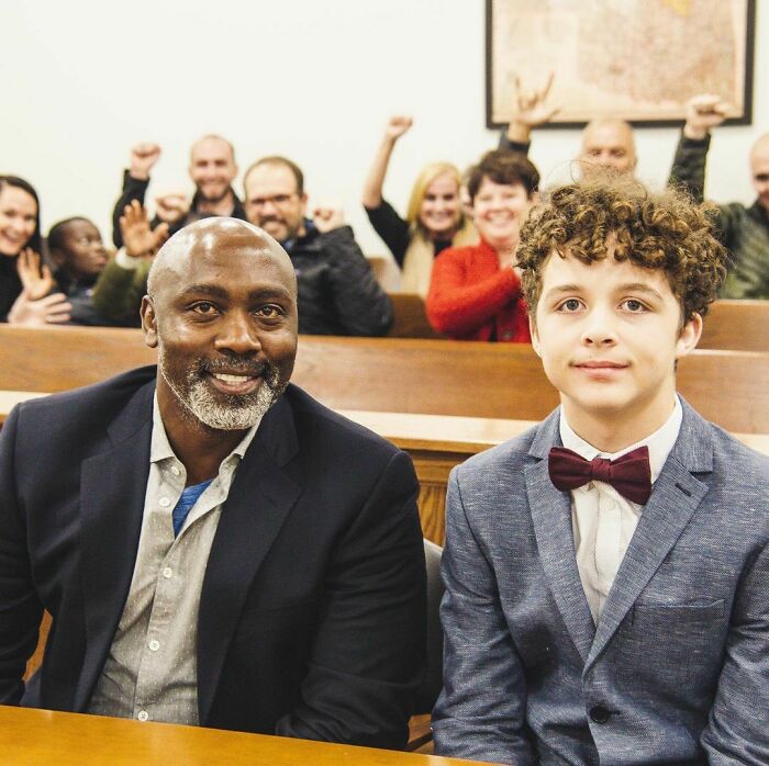 Smiling man and boy at an adoption ceremony with celebratory group in the background, highlighting wholesome adoption stories.