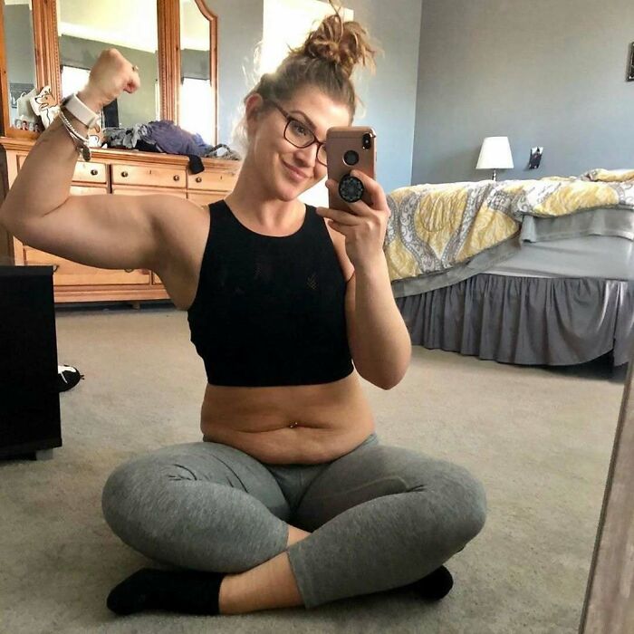 Woman taking a selfie in casual wear, flexing her arm and smiling in a cozy bedroom setting.