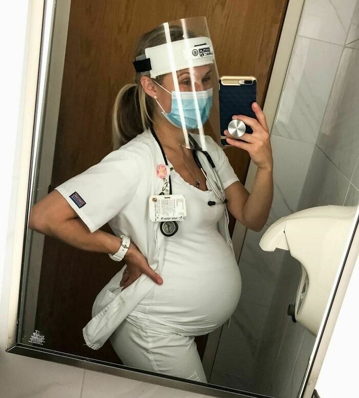 Pregnant nurse in scrubs takes a mirror selfie, wearing a mask and face shield, showcasing women in real life settings.