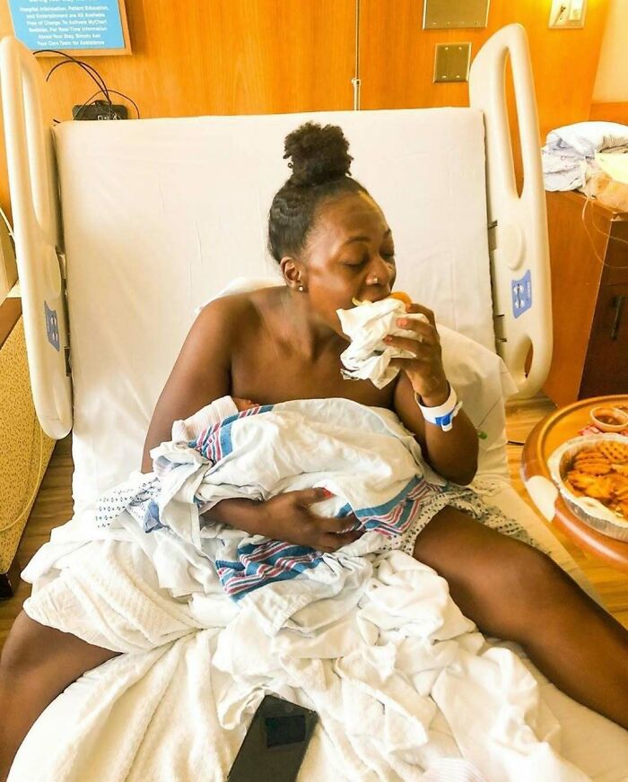 Woman on a hospital bed holding a baby, enjoying a sandwich, realistic moment in daily life.