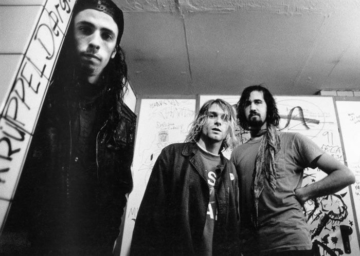 A famous trio from the band Nirvana, featuring Dave Grohl, Kurt Cobain, and Krist Novoselic standing in a gritty room with graffiti-covered walls.