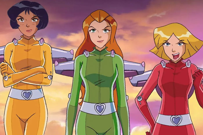 A famous trio from Totally Spies!, featuring Alex, Sam, and Clover in their yellow, green, and red spy outfits, standing confidently against a sunset sky.