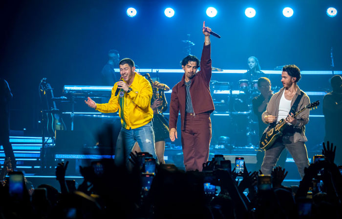 A famous trio, the Jonas Brothers, performing live on stage with Nick in a yellow jacket, Joe raising a hand, and Kevin playing guitar.