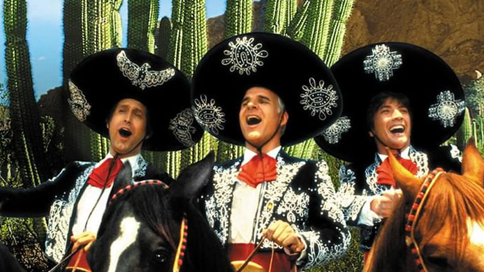 A famous trio from Three Amigos, featuring the characters Lucky Day, Dusty Bottoms, and Ned Nederlander in elaborate mariachi outfits, riding horses in a desert with cacti in the background.