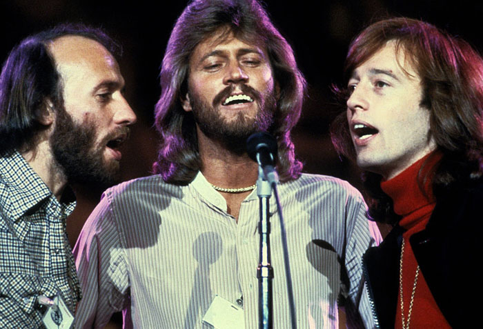 A famous trio from a music group, the Bee Gees, featuring Barry, Robin, and Maurice Gibb singing together on stage around a single microphone.