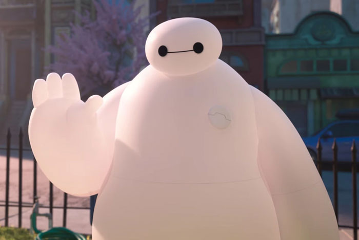 A fat cartoon character, Baymax, a white inflatable healthcare robot with a simple face, waves outdoors in a city setting with buildings and trees.