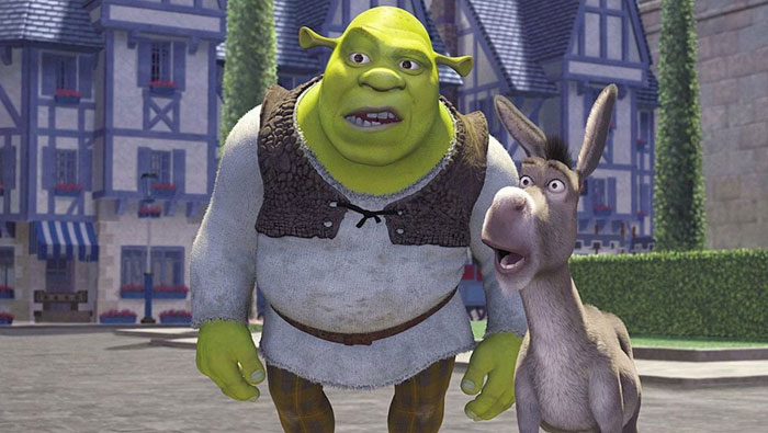 A fat cartoon character, Shrek, a green ogre in a tunic, stands beside his surprised-looking friend, Donkey, in a village with Tudor-style buildings.