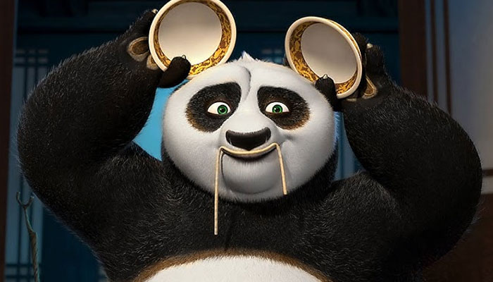 A fat cartoon character, Po the panda, playfully holds two bowls above his head with noodles hanging from his mouth, looking amused indoors.