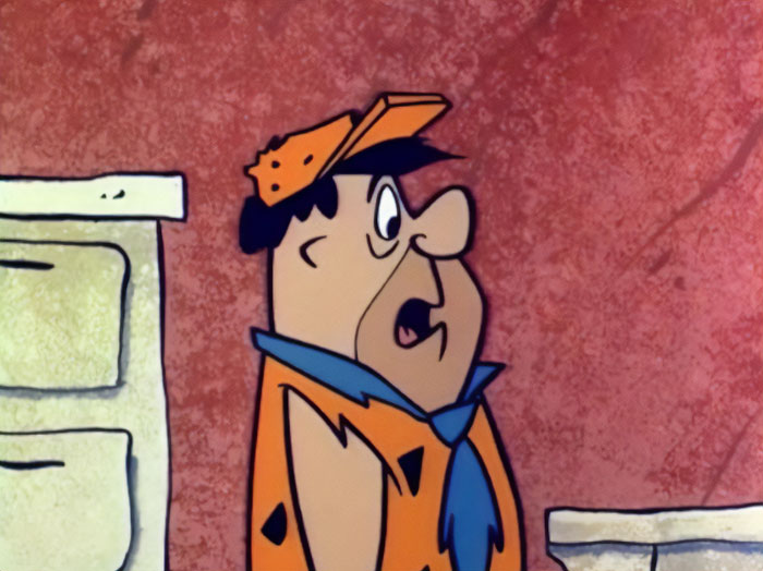  A fat cartoon character, Fred Flintstone, wearing an orange and black spotted tunic with a blue tie, looks surprised while indoors.