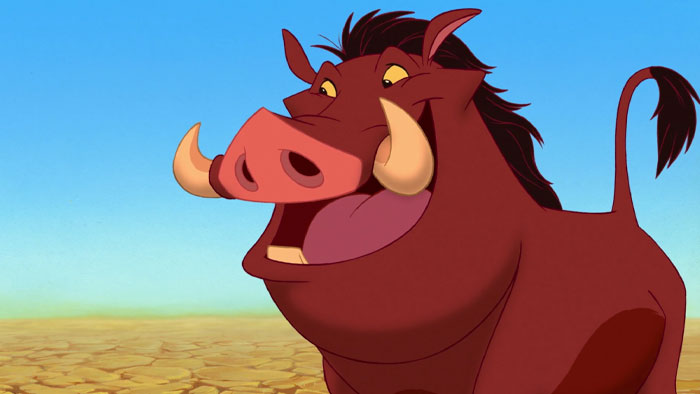 A fat cartoon character, Pumbaa the warthog, smiling widely with prominent tusks, stands on a dry, cracked landscape under a clear blue sky.