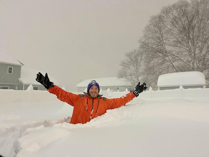 Man in orange coat and blue hat joyfully standing in deep snow, showcasing ice-cold winter scenery with snow-covered trees.