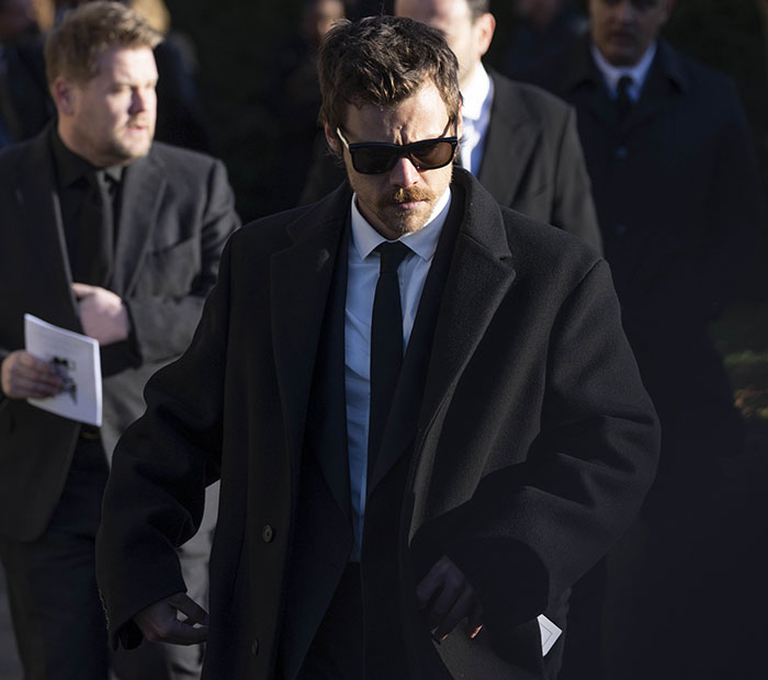 Individual in a black coat and sunglasses, walking solemnly at a formal event.