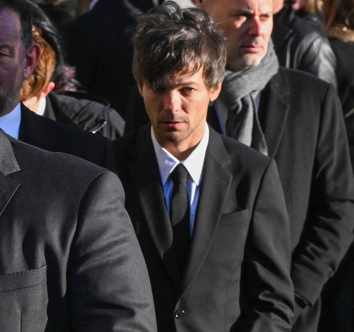 A man in a black suit and tie, looking serious, stands among a crowd, capturing a somber moment.