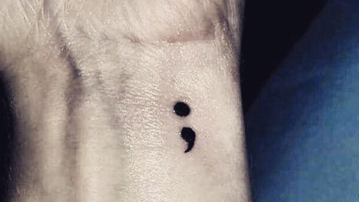 A semicolon tattoo on the wrist, symbolizing resilience and continuity, with a bold and minimalist design against a textured skin background.