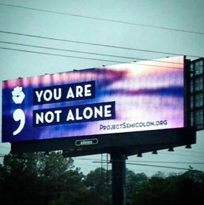 A billboard with a semicolon symbol and the message "YOU ARE NOT ALONE" by ProjectSemicolon.org, promoting mental health awareness and support.