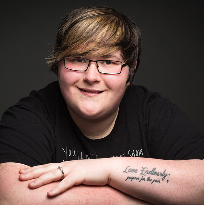 A person with glasses smiling, displaying a semicolon tattoo on their forearm that reads, "Love Endlessly, purpose for the pain," symbolizing resilience.