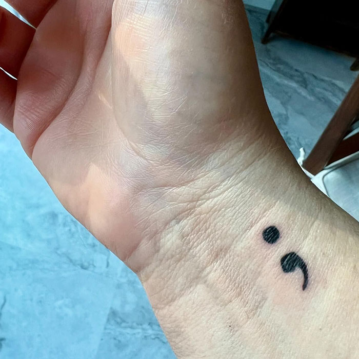 A bold semicolon tattoo on the wrist, symbolizing resilience and continuation, against a background of natural light and neutral flooring.