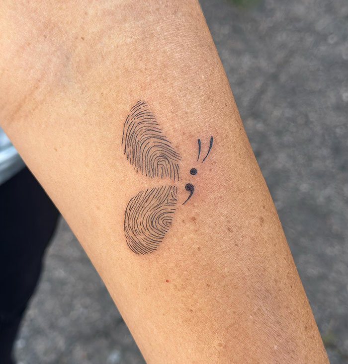 A semicolon tattoo on the forearm designed as a butterfly with fingerprint wings, symbolizing uniqueness, resilience, and transformation.