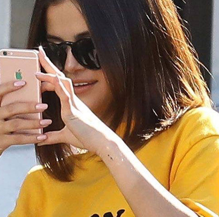 Selena Gomez wearing sunglasses and a yellow shirt, holding a smartphone with a small semicolon tattoo on her wrist, symbolizing mental health awareness.