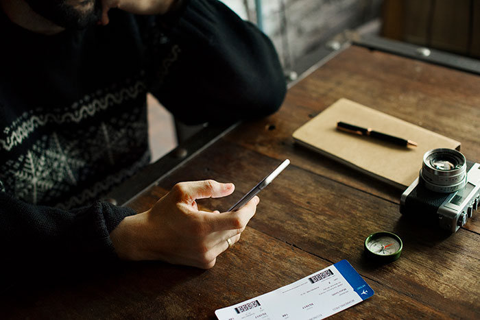 Passenger using a phone at a wooden table with tickets, notebook, camera, and compass nearby.