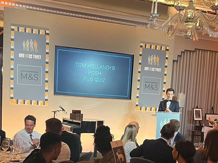 A charity event with a banner displaying "Tom Holland's Posh Pub Quiz," featuring Tom Holland speaking at a podium in a formal setting.