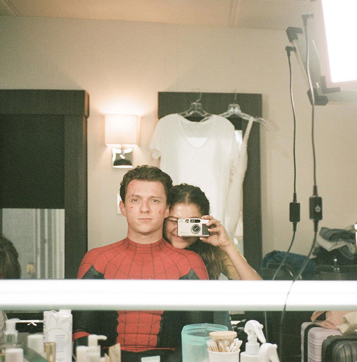 A behind-the-scenes photo showing Tom Holland dressed as Spider-Man, with Zendaya playfully holding a camera behind him, both reflected in a makeup mirror.