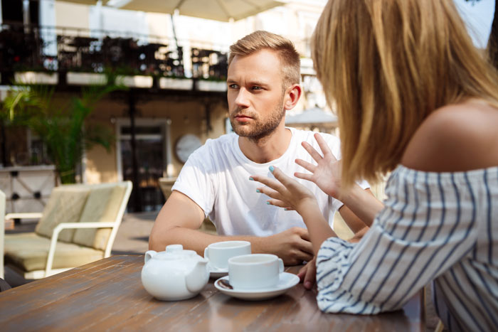 Two people at a cafe table, conversing with intense expressions, coffee cups nearby.