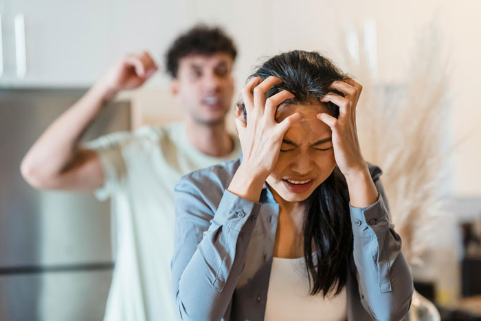 A frustrated woman holds her head while a man argues in the background, depicting family tension about a college fund.