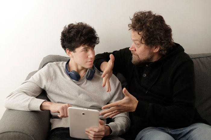 Father and son having a serious conversation on the couch about the son's future, with a tablet in the son's hand.