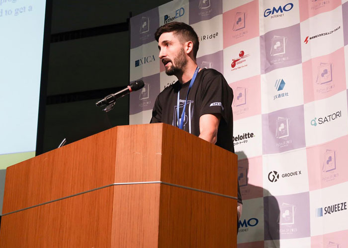 Bobbi Althoff’s husband, speaking at a podium during a conference, wearing a black shirt, against a branded backdrop.