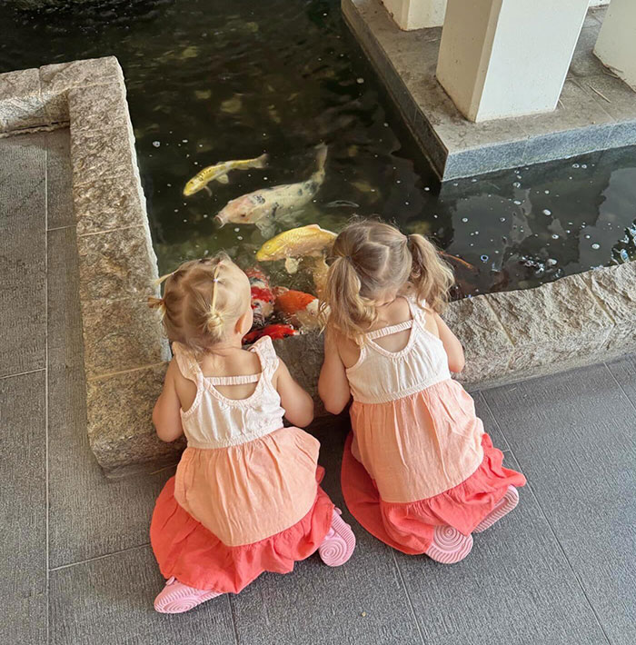 Two young children, connected to Bobbi Althoff’s husband, sitting by a koi pond, wearing matching dresses, gazing at colorful fish.