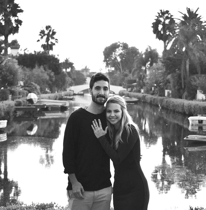 A black-and-white photo of Bobbi and Cory Althoff standing by a serene canal, smiling, surrounded by palm trees and a picturesque bridge.