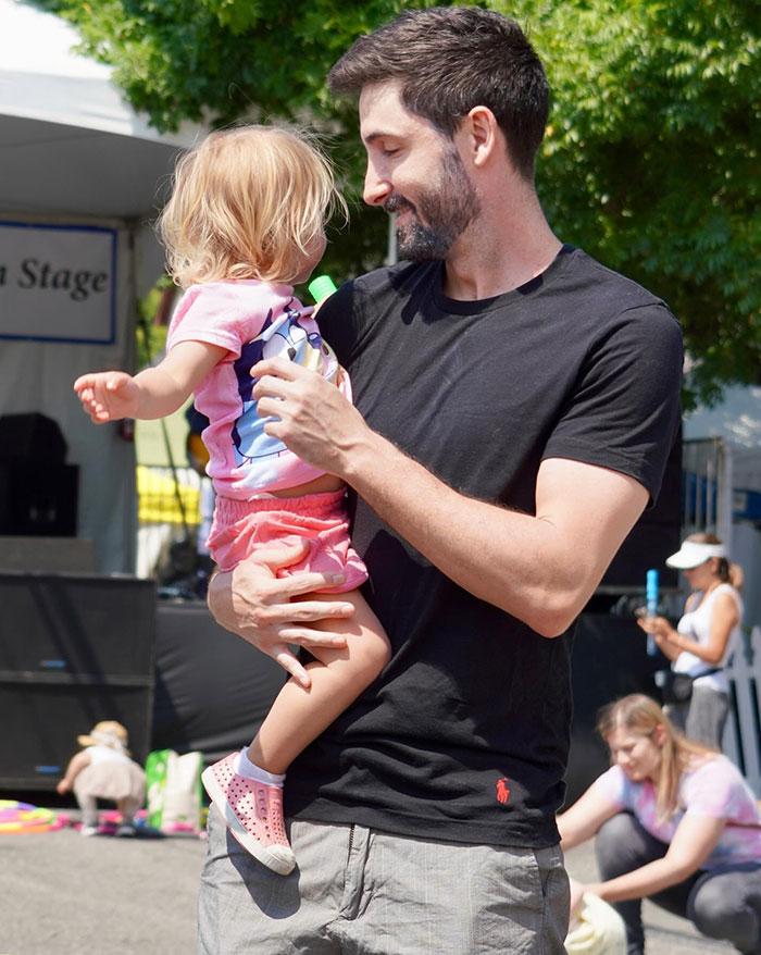 Bobbi Althoff’s husband, smiling and holding a young child at an outdoor event, with people and trees visible in the background.