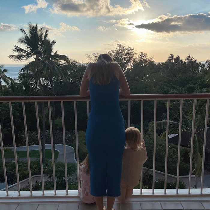 Bobbi Althoff, in a blue dress, leaning on a balcony railing with two young children at her side, overlooking a tropical sunset.