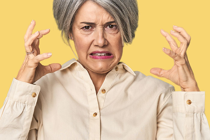 Woman with gray hair looking frustrated, wearing a beige shirt, against a yellow background, expressing discomfort.