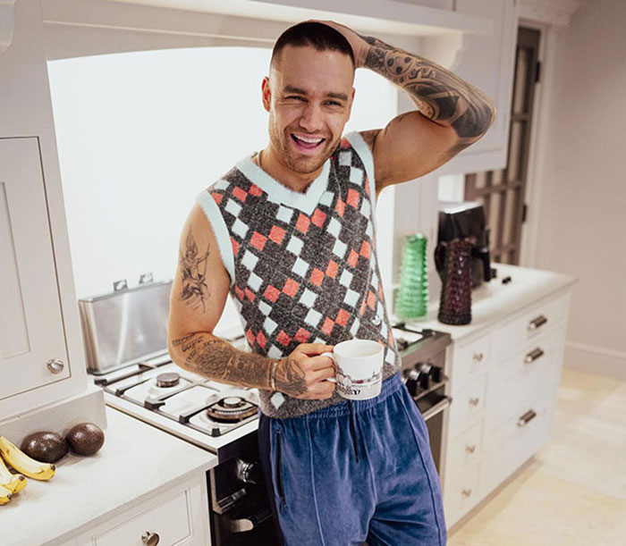 Man with tattoos in kitchen, holding a mug, smiling, and wearing a patterned vest and blue pants.